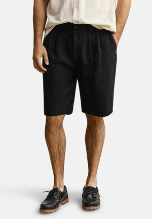 Mann trägt schwarze maßgeschneiderte Shorts, beige kurzärmeliges Hemd und schwarze Lederschuhe mit braunen Sohlen, steht mit den Händen in den Taschen.
