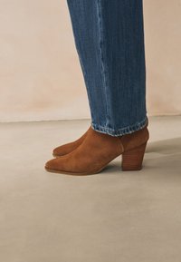 Bruine suède enkellaars met een puntige neus en een gestapelde houten hak. Gecombineerd met donkerblauwe rechte spijkerbroeken, waarbij de zoomdetails zichtbaar zijn.