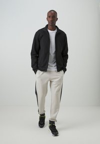 Czarna kurtka na zamek, szary t-shirt, kremowe dresy z czarnymi bocznymi panelami i czarne sneakersy z żółtymi akcentami, stojące na neutralnym tle.