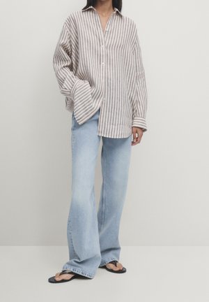 Osoba ubrana w luźną beżowo-białą koszulę w paski zapinaną na guziki, jasnoniebieskie szerokie dżinsy i czarne klapki, stojąca na jednolitym tle.