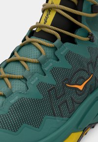 Τρέχον αθλητικό παπούτσι σε τιρκουάζ χρώμα με υφασμάτινο επάνω μέρος, πορτοκαλί λεπτομέρειες και κίτρινες διακοσμήσεις. Διαθέτει σχεδιασμό από πλεκτό δίχτυ με μοτίβο και ανθεκτικό σύστημα δέσης κορδονιών.