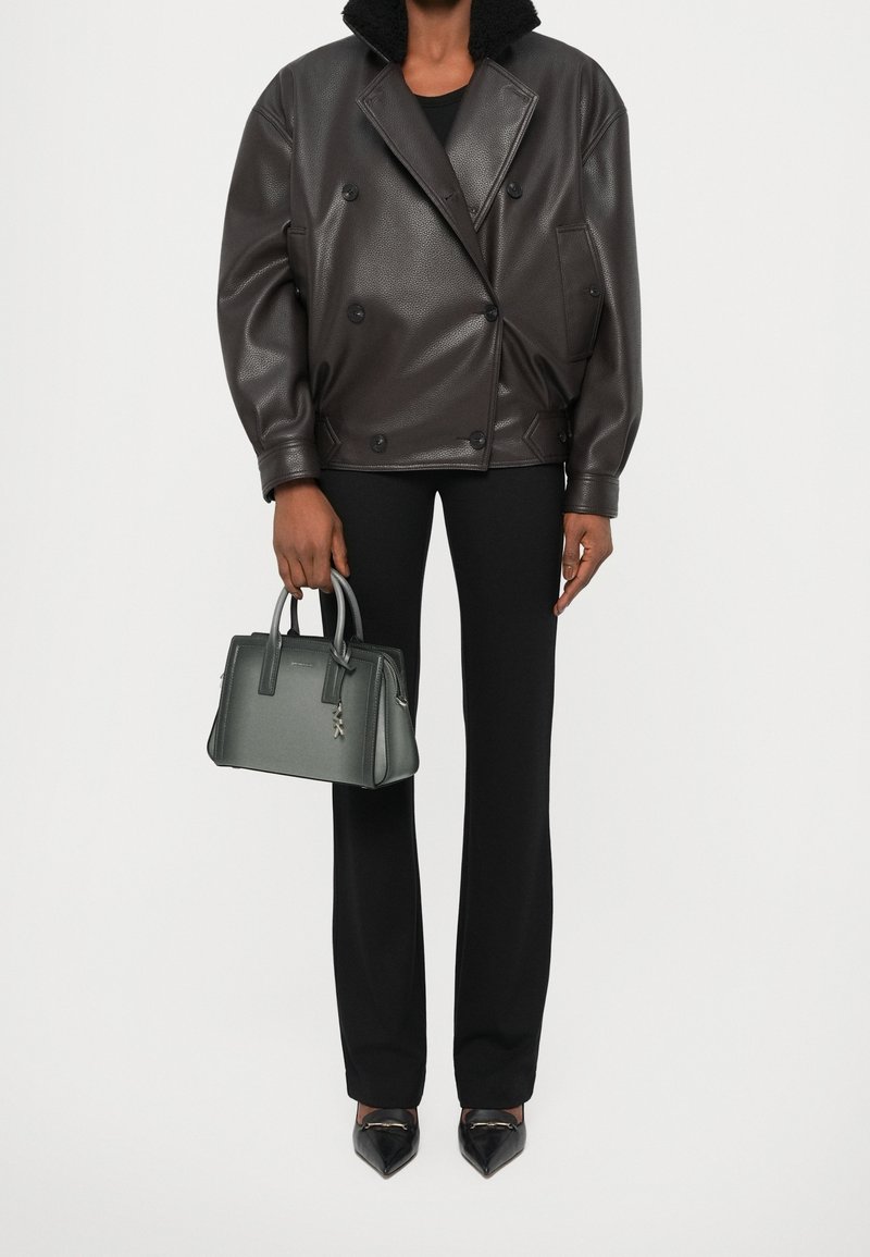 Personne portant une veste en cuir marron foncé, un pantalon noir, des chaussures noires pointues, et tenant un petit sac à main gris avec deux poignées.