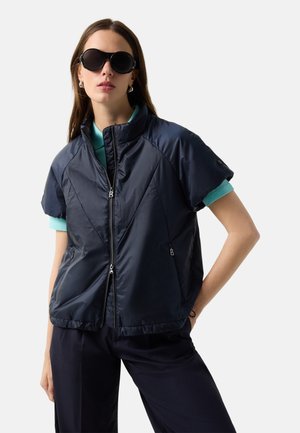 Kobieta w czarnych okularach przeciwsłonecznych, krótkim granatowym puchowym płaszczu na turkusowej koszulce i granatowych spodniach na tle jasnego, jednolitego tła.