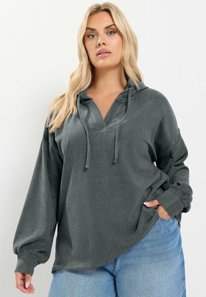 Femme aux longs cheveux blonds portant un sweat à capuche ample gris foncé avec cordons et un jean bleu clair, une main dans la poche.