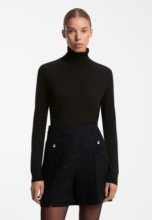 Mujer con jersey de cuello alto negro y pantalones cortos negros de talle alto con textura y detalles de botones en los bolsillos sobre fondo blanco.