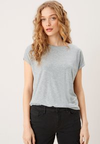Jasnoszara koszulka z krótkim rękawem o luźnym kroju i okrągłym dekoltem, wykonana z miękkiego, teksturowanego materiału. Noszona z czarnymi spodniami.