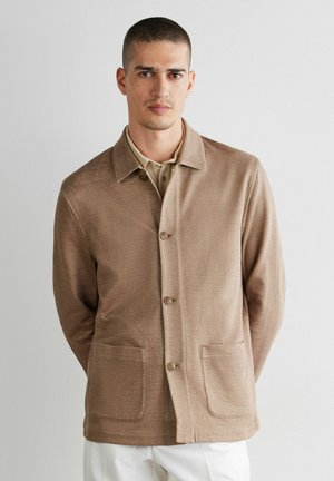 Mand med kort hår iført en beige knapjakke over en lyse skjorte, stående foran en ensfarvet, lys baggrund.