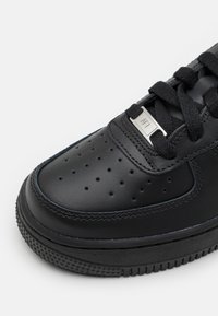 Sneaker de couro preto com perfurações na parte frontal, parte superior texturizada e atacadores pretos planos com um detalhe prateado na zona dos atacadores.