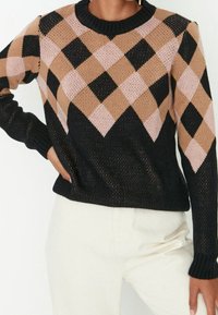 Femme portant un pull noir avec un motif losange beige et rose clair, associé à un pantalon blanc taille haute.