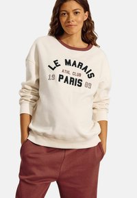 Sudadera crema con cuello burdeos y "LE MARAIS ATHL CLUB PARIS 1989" impreso en negrita negro. Combinada con pantalones de chándal burdeos.