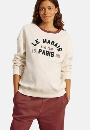 Sudadera crema con cuello burdeos y "LE MARAIS ATHL CLUB PARIS 1989" impreso en negrita negro. Combinada con pantalones de chándal burdeos.