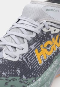 Scarpa da corsa grigia con superficie testurizzata, occhielli rinforzati e un logo giallo prominente. Presenta una suola intermedia verde chiaro con un design a motivo.