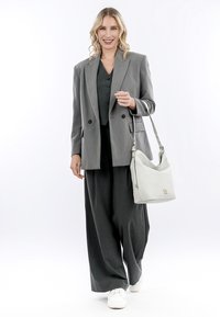 Grijze op maat gemaakte blazer over een donker vest, gecombineerd met wijde broekspijpen en witte sneakers. Houdt een lichtgrijze schoudertas vast. Glad textuur.
