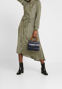 Green patterned dress with a tie waist, paired with a black leather handbag featuring an orange handle and black ankle boots.