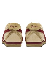 Béžové semišové tenisky s červenými akcenty, vybavené polstrovaným límcem a logem "Tiger" na patě, plochou gumovou podrážkou a detaily v překřížení.