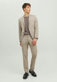 Completo a righe beige con vestibilità slim, giacca monopetto e pantaloni abbinati. Camicia grigia a girocollo sotto, abbinata a scarpe nere.