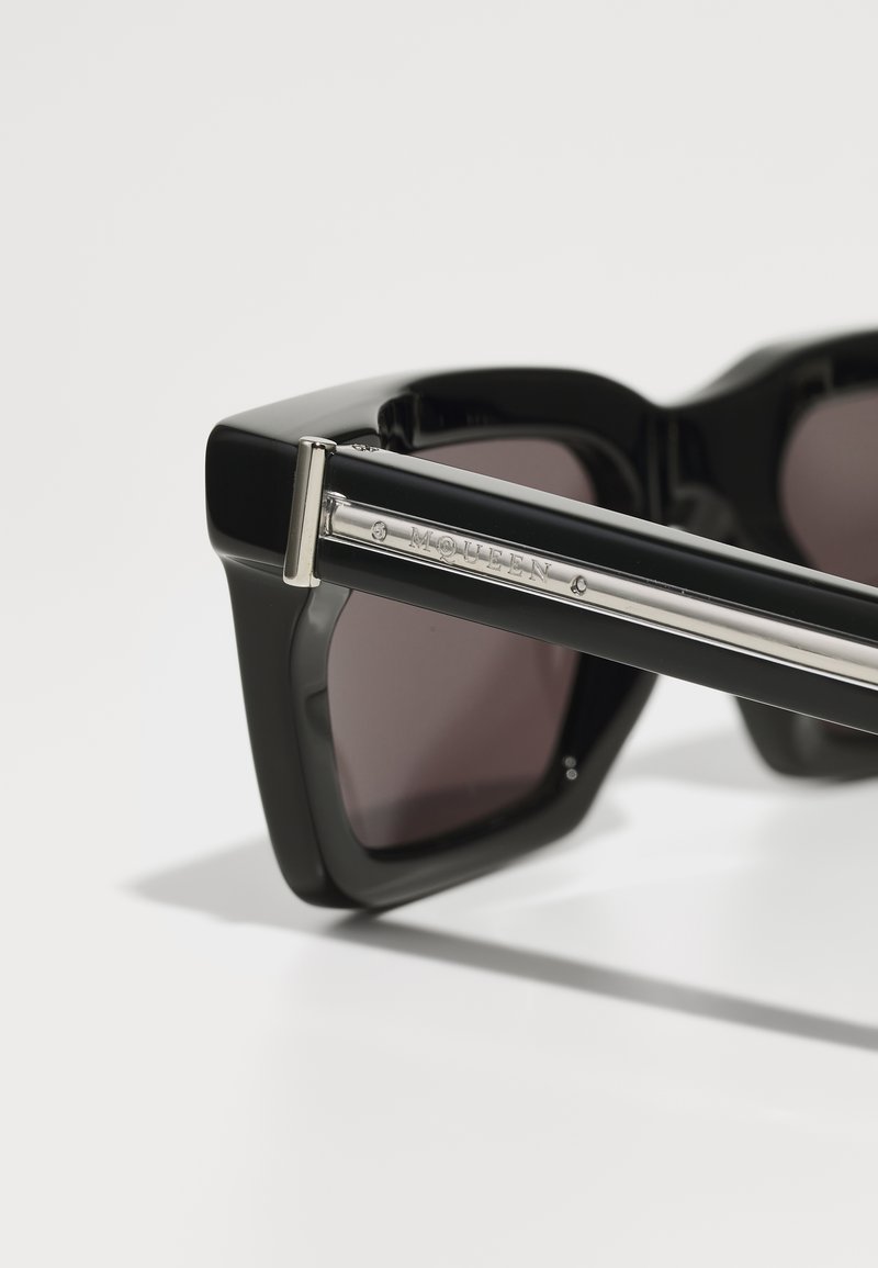 Lunettes de soleil noires avec une large monture angulaire, des verres teintés et un accent argenté sur la branche avec un logo en relief.