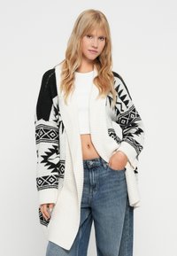 Dolga cardigan v črno-beli pletenini, ki ima abstraktni vzorec in rebrast ovratnik, v kombinaciji z modrimi hlačami z visokim pasom.