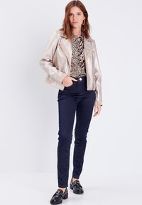Veste de biker beige métallique avec une fermeture éclair à l'avant et des accents contrastants sur les épaules, associée à un blouse à imprimé léopard et un jean skinny noir.