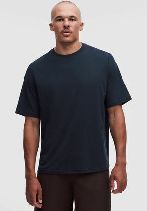 Uomo con capelli corti e baffi che indossa una maglietta semplice blu scuro a maniche corte e pantaloni scuri su uno sfondo chiaro.