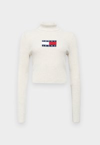 Maglione a collo alto a coste crema con maniche lunghe. Presenta un'etichetta rettangolare con "TOMMY JEANS" nei colori navy, rosso e bianco.