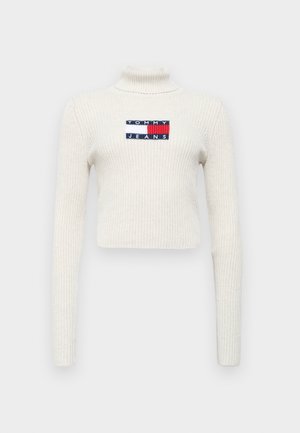 Maglione a collo alto a coste crema con maniche lunghe. Presenta un'etichetta rettangolare con "TOMMY JEANS" nei colori navy, rosso e bianco.