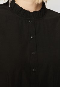 Μαύρη μπλούζα με ριχτό γιακά, που διαθέτει έξι μαύρα κουμπιά μπροστά. Λείο ύφασμα με ματ φινίρισμα.