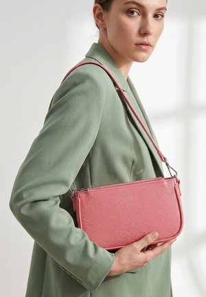 Vrouw in een saliegroene blazer die een roze getextureerde leren schoudertas met zilveren details aan haar zijde vasthoudt.