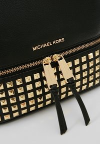 Sac Michael Kors noir avec fermeture éclair dorée, deux curseurs et clous carrés dorés sur la partie inférieure.