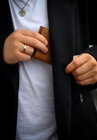Brown leather wallet being pulled from a black coat pocket by a hand wearing a silver ring. The person is wearing a white t-shirt underneath.