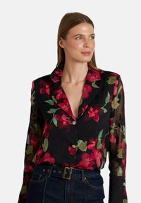 Blusa negra con estampado floral que presenta flores rojas y verdes, mangas largas transparentes y un cuello con botones decorativos.