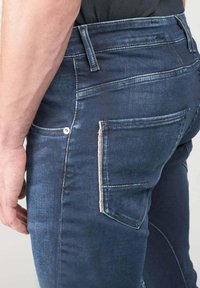 Jeans de mezclilla azul oscuro con corte ajustado, con costuras blancas, dos bolsillos traseros y un detalle de botón de metal en el bolsillo.