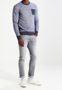 Felpa blu navy con un aspetto testurizzato, caratterizzata da una tasca sul petto blu scuro, abbinata a jeans grigi chiari e sneakers bianche.