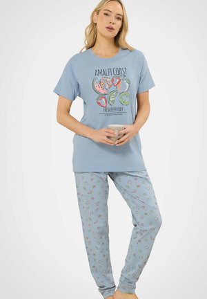 Kobieta w niebieskim komplecie piżamowym z nadrukiem owoców na koszuli i spodniach, trzymająca beżowy kubek obiema rękami.