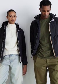 Zwei Personen tragen Jacken; eine in Schwarz mit weißen Akzenten, die andere in Olivgrün. Das weibliche Model hat ein helles, geripptes Oberteil und helle Jeans an.