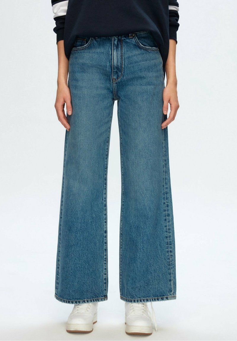 Personne portant un jean bleu taille haute à jambes larges et des baskets blanches avec un haut à manches longues foncé sur fond blanc.