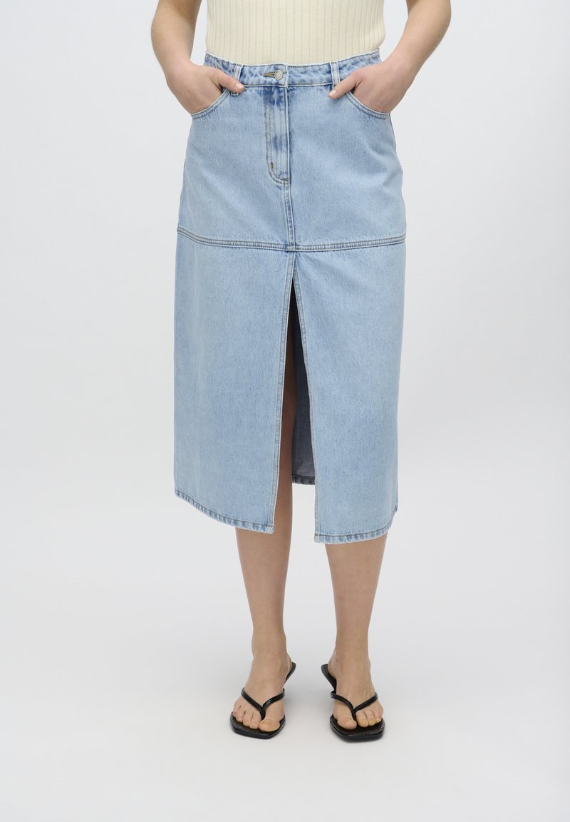 Vaaleansininen denim-midi-hame, jossa on etuaukko, sivutaskut ja erottuvat tikkaukset saumoissa. Yhdistetty mustiin sandaaleihin.