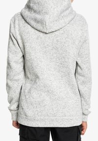 Persona che indossa una felpa con cappuccio grigia chiara e maculata, mostrata di spalle, con pantaloni neri e mani rilassate ai lati.