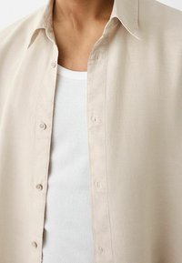 Beżowa koszula z długim rękawem wykonana z lekkiego materiału, z guzikami, miękką teksturą i kołnierzykiem, noszona na białym topie na ramiączkach.