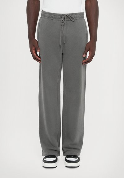 Un pantalon de survêtement gris avec une taille à cordon de serrage, des poches latérales et une coupe large, associé à des baskets noires et blanches. Texture lisse.