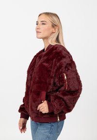 Giacca bomber in pelliccia sintetica bordeaux con polsini e orlo a coste, dotata di una tasca con zip sulla manica e una superficie morbida e texturizzata.