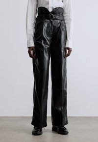 Svarta läderbyxor med hög midja och vida ben. Har ett texturerat bälte med ett runt spänne. Slät, glansig finish.