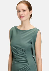 Grünes Kleid mit glatter Textur, taillierter Passform, weitem Ausschnitt und gerafftem Detail an der Seite. Ärmellos mit verlängertem Schulterstoff.