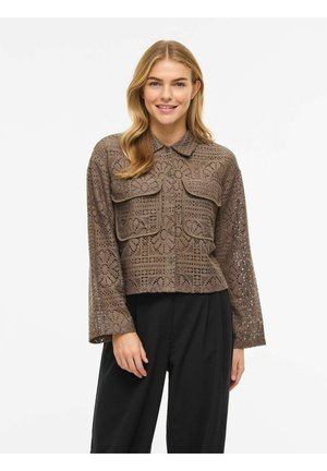 Kvinde iført en brun hæklet skjorte med knapper og store lommer samt sorte bukser, smilende foran en ensfarvet hvid baggrund.