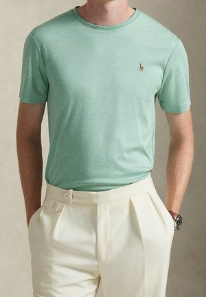 Man draagt een lichtgroen T-shirt met ronde hals met klein paardenlogo en crème kleurige geplooide broek met hoge taille en dubbele knoop detail.