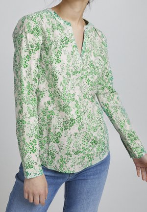 Vrouw draagt een blouse met lange mouwen en een groene bloemenprint, gecombineerd met een blauwe spijkerbroek, staand tegen een effen lichtgrijze achtergrond.