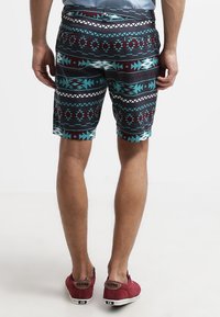 Muž oblečený v geometrických šortkách s vzorem v modré, černé a vínové barvě, světle modré košili a červených teniskách, stojící zády k divákovi.