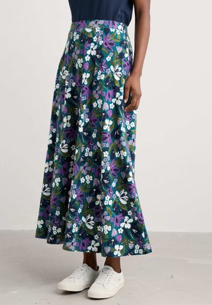 Femme portant une longue jupe fluide à fleurs vertes, violettes et blanches, associée à des baskets blanches et un haut bleu marine.