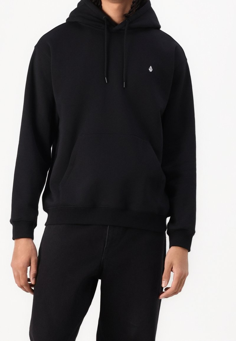 Personne portant un sweat à capuche noir uni avec un petit logo blanc sur la poitrine et un pantalon noir, debout sur un fond blanc.