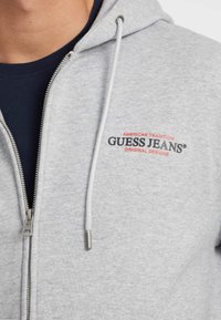 Sudadera gris con cremallera hecha de una tela suave, con capucha ajustable. Presenta un logo impreso de "GUESS JEANS" en rojo y negro en el pecho.